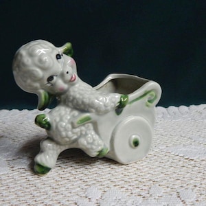 May include: A white ceramic figurine of a lamb pulling a small cart. The lamb has curly textured fur, pink cheeks, and green painted accents on its ears, hooves, and the cart wheels. The cart features a hollow opening at the top.