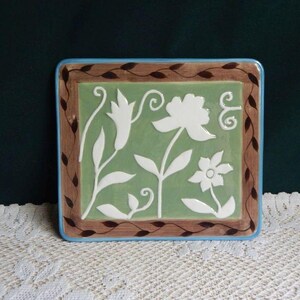 May include: Square ceramic decorative tile with a floral design. The tile features a light green center with white flowers and leaves. A brown border with dark brown leaves surrounds the green center. The tile has a blue edge.