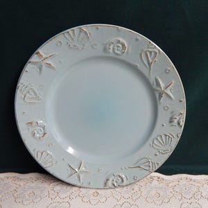 May include: A light blue ceramic plate with embossed seashell and starfish designs around the rim. The plate has a slightly distressed, rustic appearance, with a diameter of approximately 10 inches.
