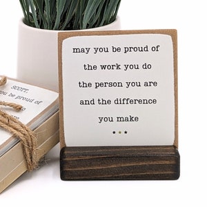 May include: A set of small cards with inspirational quotes. The card shown reads "may you be proud of the work you do the person you are and the difference you make". The cards are displayed on a wooden stand.