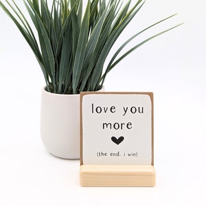 Può includere: Un piccolo cartello di legno con le parole "love you more" e un simbolo a forma di cuore, appoggiato su un supporto di legno. Il cartello è davanti a un vaso bianco con erba verde.