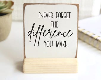 May include: A square white wooden sign with dark brown edges, featuring the black text "NEVER FORGET THE difference YOU MAKE" in a mix of block and script fonts, resting in a light-colored natural wood block stand.
