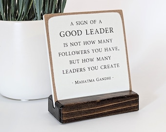 Peut inclure: Une pancarte blanche avec un cadre en bois brun et un support noir. La pancarte indique "A sign of a good leader is not how many followers you have, but how many leaders you create - Mahatma Gandhi -"