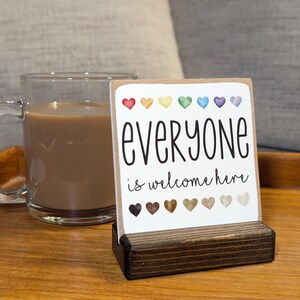 May include: A wooden sign with the text "everyone is welcome here" in black script. The sign is decorated with a row of rainbow colored hearts and a row of brown hearts. The sign is sitting on a wooden stand.