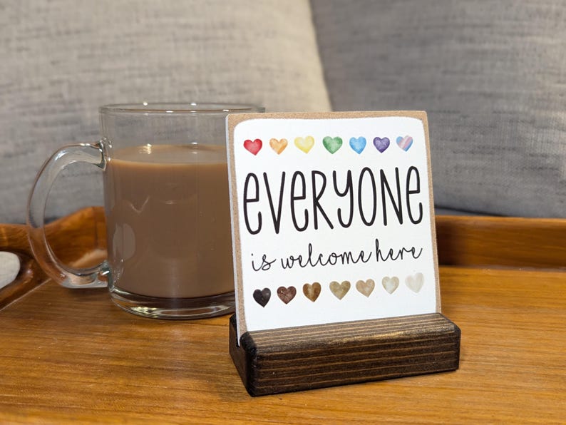 May include: A wooden sign with the text "everyone is welcome here" in black script. The sign is decorated with a row of rainbow colored hearts and a row of brown hearts. The sign is sitting on a wooden stand.