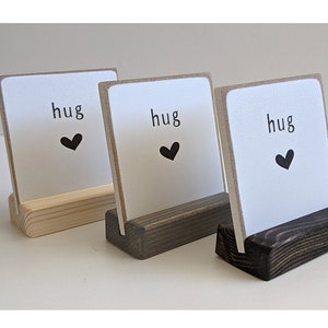 May include: Three square white signs with the word hug and a black heart icon printed on each. Each sign is held upright by a rectangular wooden base, with the bases finished in light, medium, and dark brown wood tones respectively.