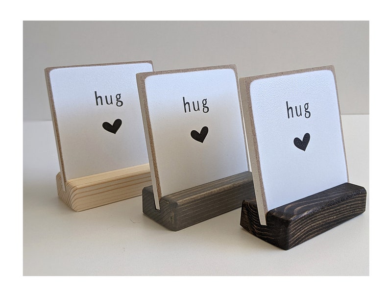 May include: Three square white signs with the word hug and a black heart icon printed on each. Each sign is held upright by a rectangular wooden base, with the bases finished in light, medium, and dark brown wood tones respectively.