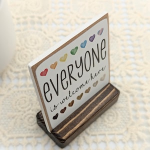 May include: A wooden sign with the text "Everyone is welcome here" in black lettering. The sign is decorated with colorful hearts and is displayed on a wooden stand.