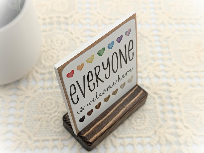 May include: A wooden sign with the text "Everyone is welcome here" in black lettering. The sign is decorated with colorful hearts and is displayed on a wooden stand.