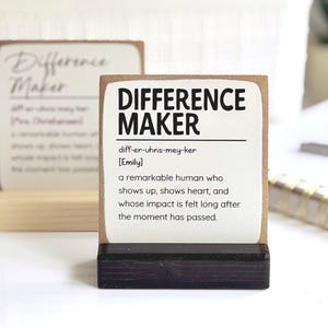 May include: A rectangular wooden sign with a black base featuring the text "DIFFERENCE MAKER" in bold black letters, followed by the phonetic spelling, the name "Emily," and a definition of a remarkable human who shows heart.