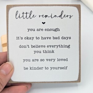 May include: A square wooden sign with a white face and brown edges. The text reads: little reminders, you are enough, it's okay to have bad days, don't believe everything you think, you are so very loved, be kinder to yourself.