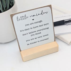 May include: A square white sign with brown edges held by a light wood stand. The text reads: little reminders, you are enough, it's okay to have bad days, don't believe everything you think, you are so very loved, be kinder to yourself.