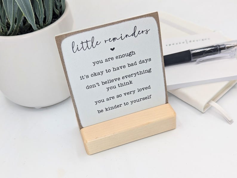 May include: A square white sign with brown edges held by a light wood stand. The text reads: little reminders, you are enough, it's okay to have bad days, don't believe everything you think, you are so very loved, be kinder to yourself.