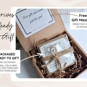 May include: A brown cardboard gift box filled with shredded paper filler. Inside, a small square item is wrapped in a clear cellophane bag and tied with a brown twine bow. A white card with the text "Difference Maker" is visible inside the bag.