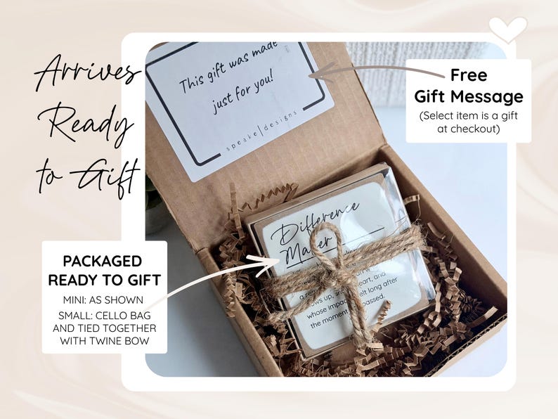 May include: A brown cardboard gift box filled with shredded paper filler. Inside, a small square item is wrapped in a clear cellophane bag and tied with a brown twine bow. A white card with the text "Difference Maker" is visible inside the bag.