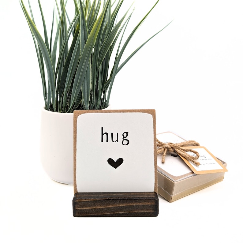 以下が含まれることがあります： 白いセラミックタイルに「hug」という言葉と黒いハートが印刷されています。タイルは濃い茶色の木製スタンドの上に置かれています。