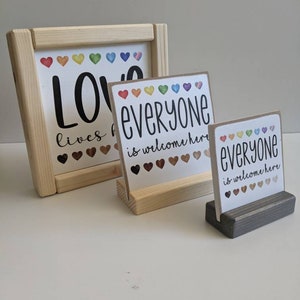 May include: Three wooden signs with the text "Love lives here", "Everyone is welcome here", and "Everyone is welcome here". The signs have a rainbow of hearts on them.