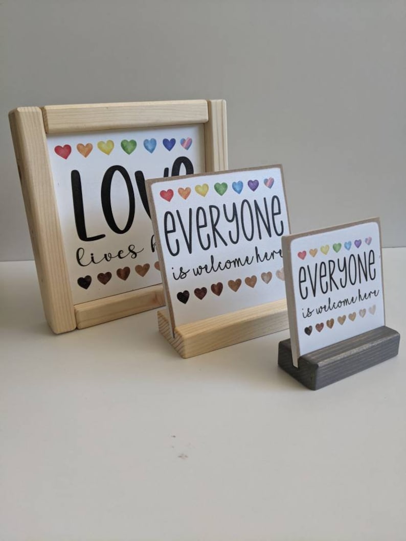 May include: Three wooden signs with the text "Love lives here", "Everyone is welcome here", and "Everyone is welcome here". The signs have a rainbow of hearts on them.