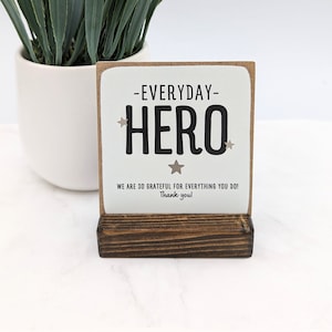 May include: White wooden sign with the text "Everyday Hero" in black with gold stars. The sign is on a small wooden stand. The text below the main text reads "We are so grateful for everything you do! Thank you!"