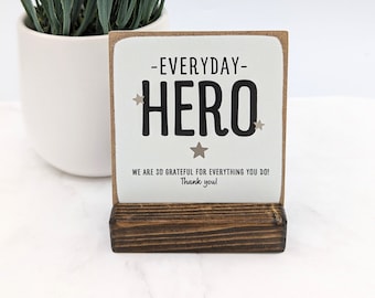 May include: White wooden sign with the text "Everyday Hero" in black with gold stars. The sign is on a small wooden stand. The text below the main text reads "We are so grateful for everything you do! Thank you!"