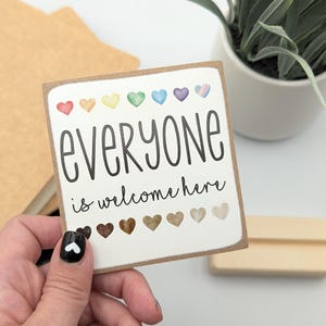 May include: A white sign with the text "everyone is welcome here" in black lettering. The sign is decorated with a row of rainbow-colored hearts at the top and a row of brown hearts at the bottom.