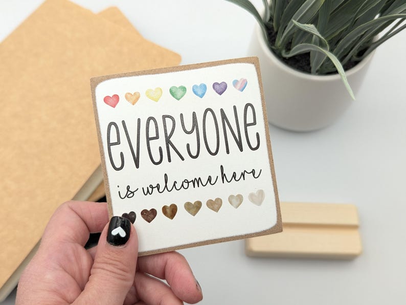May include: A white sign with the text "everyone is welcome here" in black lettering. The sign is decorated with a row of rainbow-colored hearts at the top and a row of brown hearts at the bottom.