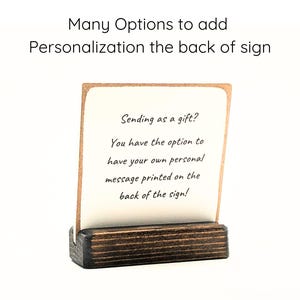 May include: A white card with a message about adding personalization to the back of a sign. The card is sitting on a small wooden stand. The text on the card reads: "Sending as a gift? You have the option to have your own personal message printed on the back of the sign!"