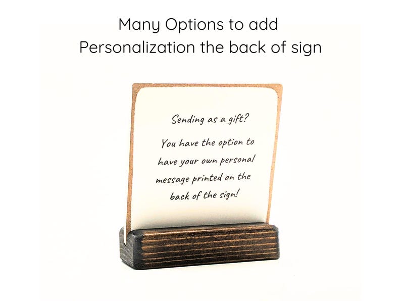 May include: A white card with a message about adding personalization to the back of a sign. The card is sitting on a small wooden stand. The text on the card reads: "Sending as a gift? You have the option to have your own personal message printed on the back of the sign!"