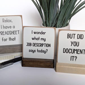 May include: Three white wooden signs with black text on a white background. The signs have the following text: "Relax, I have a SPREADSHEET for that", "I wonder what my JOB DESCRIPTION says today?", and "BUT DID YOU DOCUMENT IT?"