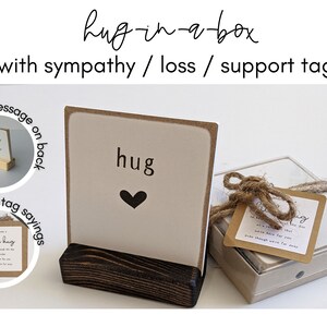 以下が含まれることがあります： 白いカードに黒いハートと「hug」と書かれた小さな木製の箱。箱は紐で結ばれており、「Sending you a little hug...」と書かれたタグが付いています。箱は白い背景の上に置かれています。