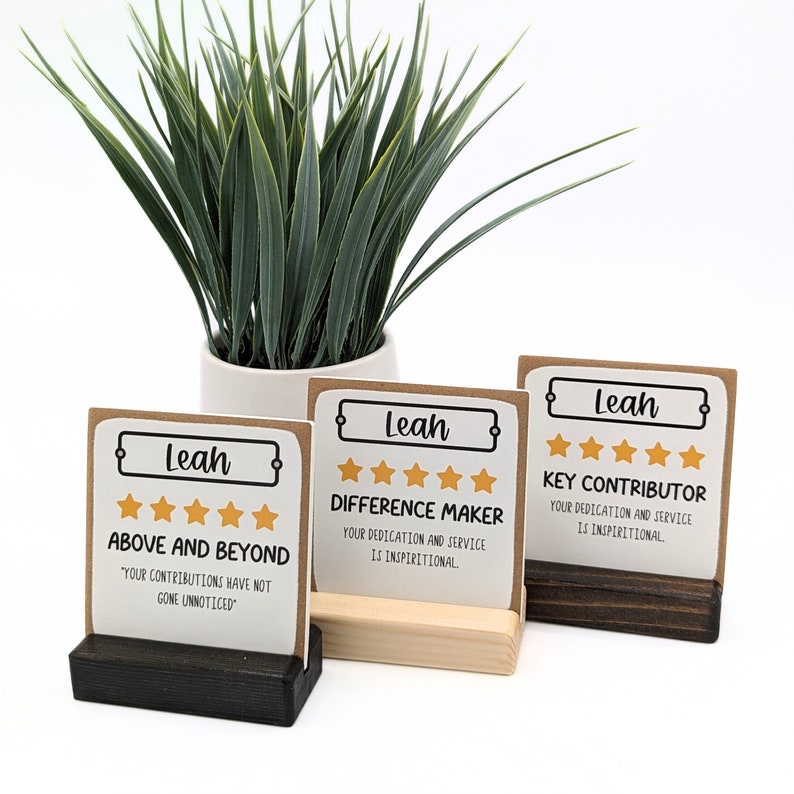 May include: Three brown card stock signs with black borders and gold stars. The signs have the name "Leah" and different phrases: "Above and Beyond", "Difference Maker", and "Key Contributor". The signs are on small wooden stands.