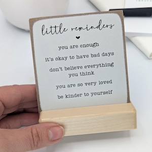 May include: A square wooden sign with a light grey face and a natural wood base. The text reads: little reminders, you are enough, it's okay to have bad days, don't believe everything you think, you are so very loved, be kinder to yourself.