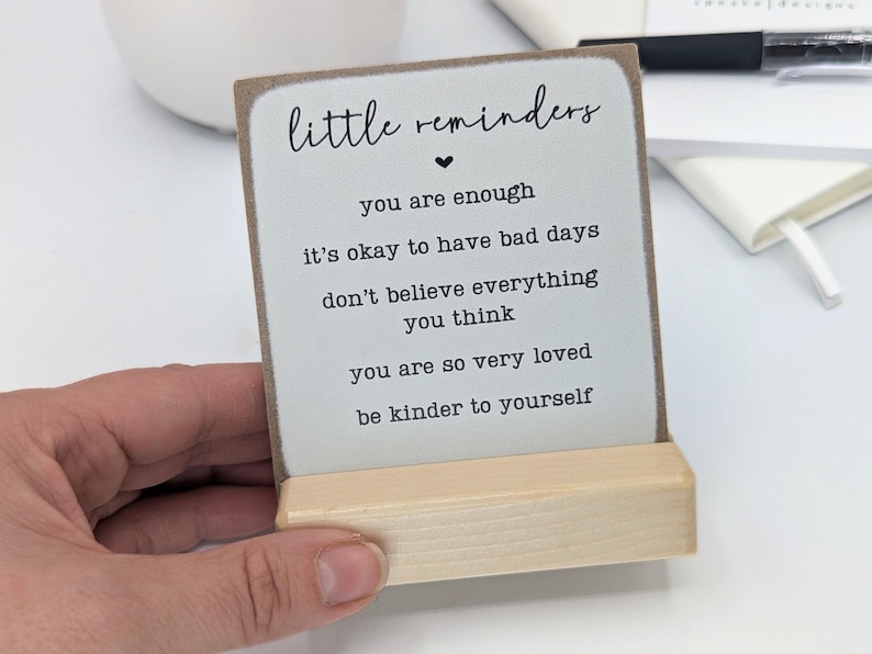 May include: A square wooden sign with a light grey face and a natural wood base. The text reads: little reminders, you are enough, it's okay to have bad days, don't believe everything you think, you are so very loved, be kinder to yourself.