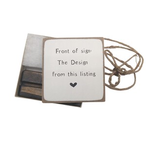 May include: A white sign with brown trim reads "Front of sign: The Design from this listing" with a small black heart below the text. The sign is in a box with a brown wooden stand and a piece of twine.