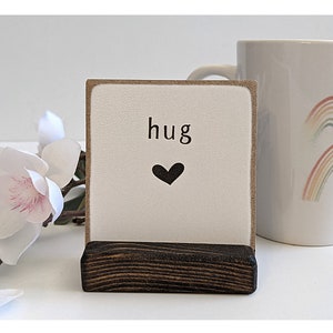 以下が含まれることがあります： 「hug」という言葉と黒いハートが印刷された小さな木製の看板。看板は小さな木製のスタンドの上に立っています。