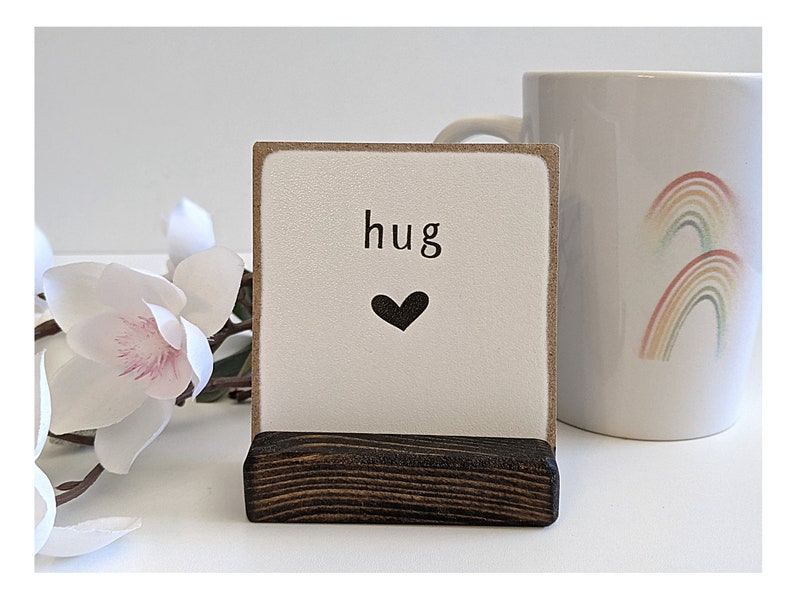以下が含まれることがあります： 「hug」という言葉と黒いハートが印刷された小さな木製の看板。看板は小さな木製のスタンドの上に立っています。