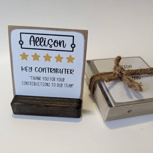 May include: A brown card with a white background and a black frame around the text "Allison" with five gold stars below. The text "Key Contributor" is below the stars. The text "Thank you for your contributions to our team" is below the text "Key Contributor". The card is on a brown wooden stand.