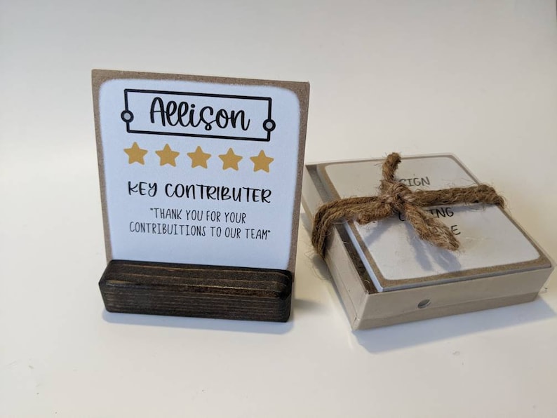 May include: A brown card with a white background and a black frame around the text "Allison" with five gold stars below. The text "Key Contributor" is below the stars. The text "Thank you for your contributions to our team" is below the text "Key Contributor". The card is on a brown wooden stand.