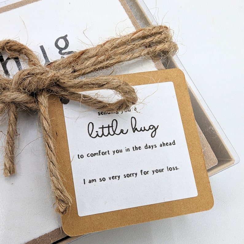 以下が含まれることがあります： 小さな茶色のカードに「little hug」という文字と「to comfort you in the days ahead I am so very sorry for your loss.」というメッセージが書かれています。カードは紐で結ばれています。