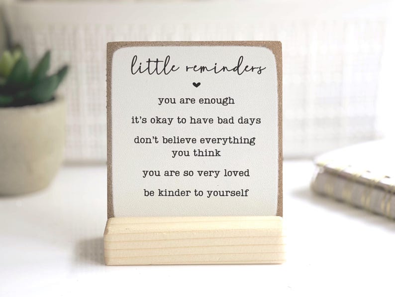 May include: A square wooden sign with a white face and brown edges, held upright by a light-coloured wooden base. The sign features the black text: little reminders, you are enough, it's okay to have bad days, don't believe everything you think, you are so very loved, be kinder to yourself.