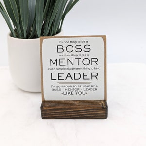 May include: A white wooden sign with a brown wooden stand. The sign reads "It's one thing to be a BOSS, another thing to be a MENTOR, but a completely different thing to be a LEADER. I'M SO PROUD TO BE LEAD BY A BOSS - MENTOR - LEADER *LIKE YOU*"
