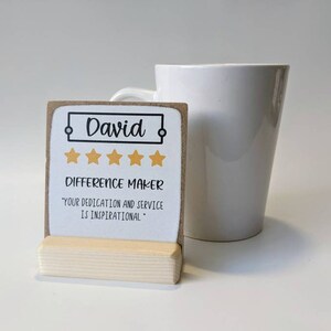 May include: A brown and white card with the name "David" in a black frame, five gold stars, and the text "Difference Maker" and "Your dedication and service is inspirational." The card is on a small wooden stand.