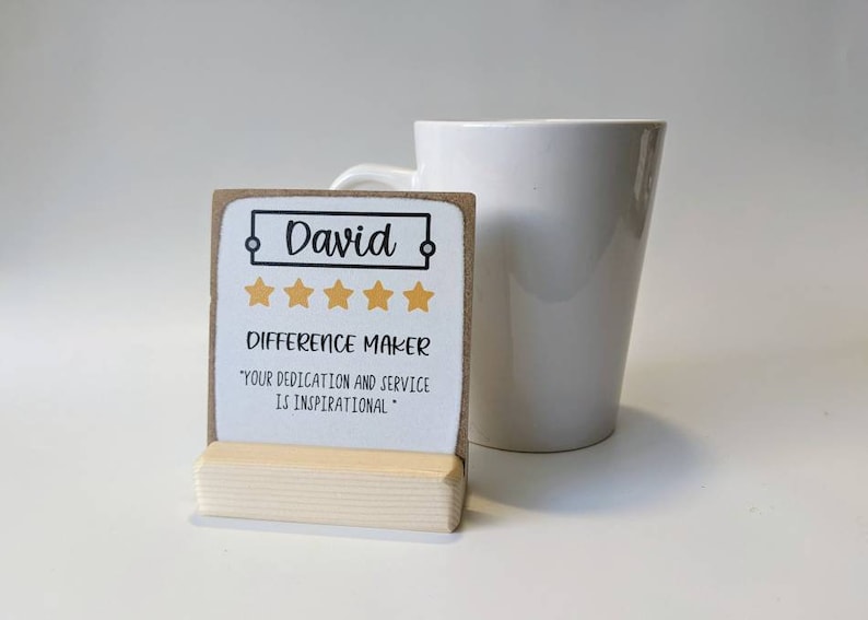 May include: A brown and white card with the name "David" in a black frame, five gold stars, and the text "Difference Maker" and "Your dedication and service is inspirational." The card is on a small wooden stand.