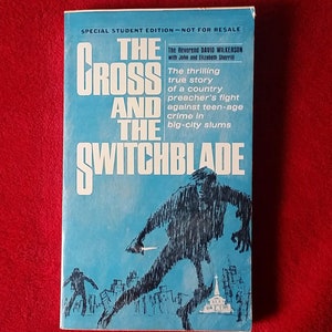 May include: A blue book cover with the title "The Cross and the Switchblade" in white text. The book cover features a black and white illustration of a person holding a switchblade in a city setting.