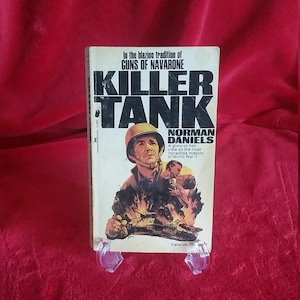 May include: A paperback book cover with a red, white, and blue color scheme. The title is "Killer Tank" in large, bold, black letters. The author is Norman Daniels. The cover features a soldier in a military uniform holding a gun, with a tank in the background. The text on the cover reads "In the blazing tradition of Guns of Navarone. A glory-or-hell crew on the most incredible mission of World War II."
