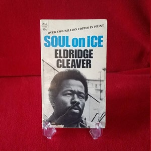 May include: A paperback book titled "Soul on Ice" by Eldridge Cleaver. The cover features a black and white portrait of the author. The book is displayed on a red surface.