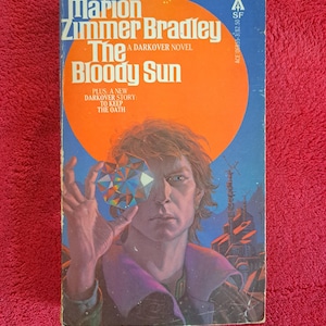 May include: A book cover with a blue background and a large orange sun. The title of the book is "The Bloody Sun" by Marion Zimmer Bradley. The book is a Darkover novel.