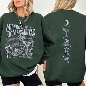 May include: Dark green sweatshirt featuring the text "Midnight Margaritas" and a cocktail illustration. The front design includes a moon, cocktail glass, and other graphics. The back features a moon, cocktail, frog, coffee pot, and mushroom.