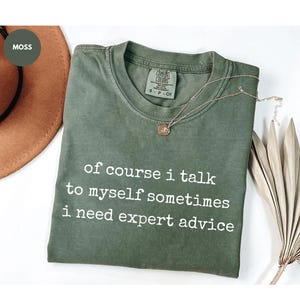 May include: Moss green t-shirt with the text "of course i talk to myself sometimes i need expert advice" in white. The shirt has a crew neck and short sleeves. A gold necklace with a pendant is draped on the shirt.