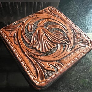 May include: A square leather case featuring intricate hand-tooled floral scrollwork patterns. The surface displays a rich tan finish with dark brown recessed areas, accented by white stitching along the edges of the leather construction.